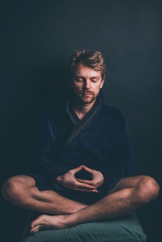 Personne assise en tailleur sur un coussin, mains devant le chakra du cœur en position de mudra, vêtements sombres, arrière-plan sombre.