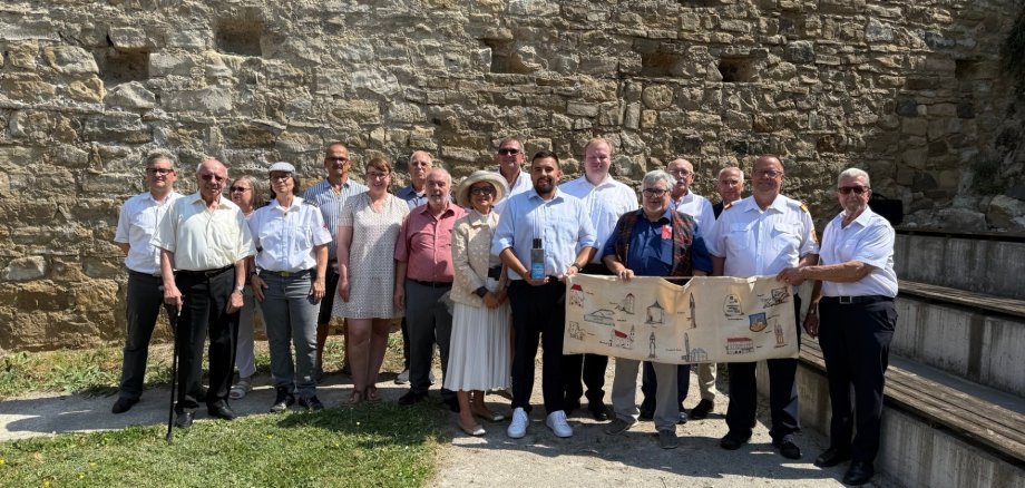 Un groupe de 14 personnes se tient devant un mur de pierre. Certains tiennent un grand papier plié avec des dessins. Les vêtements varient entre le formel et le décontracté.