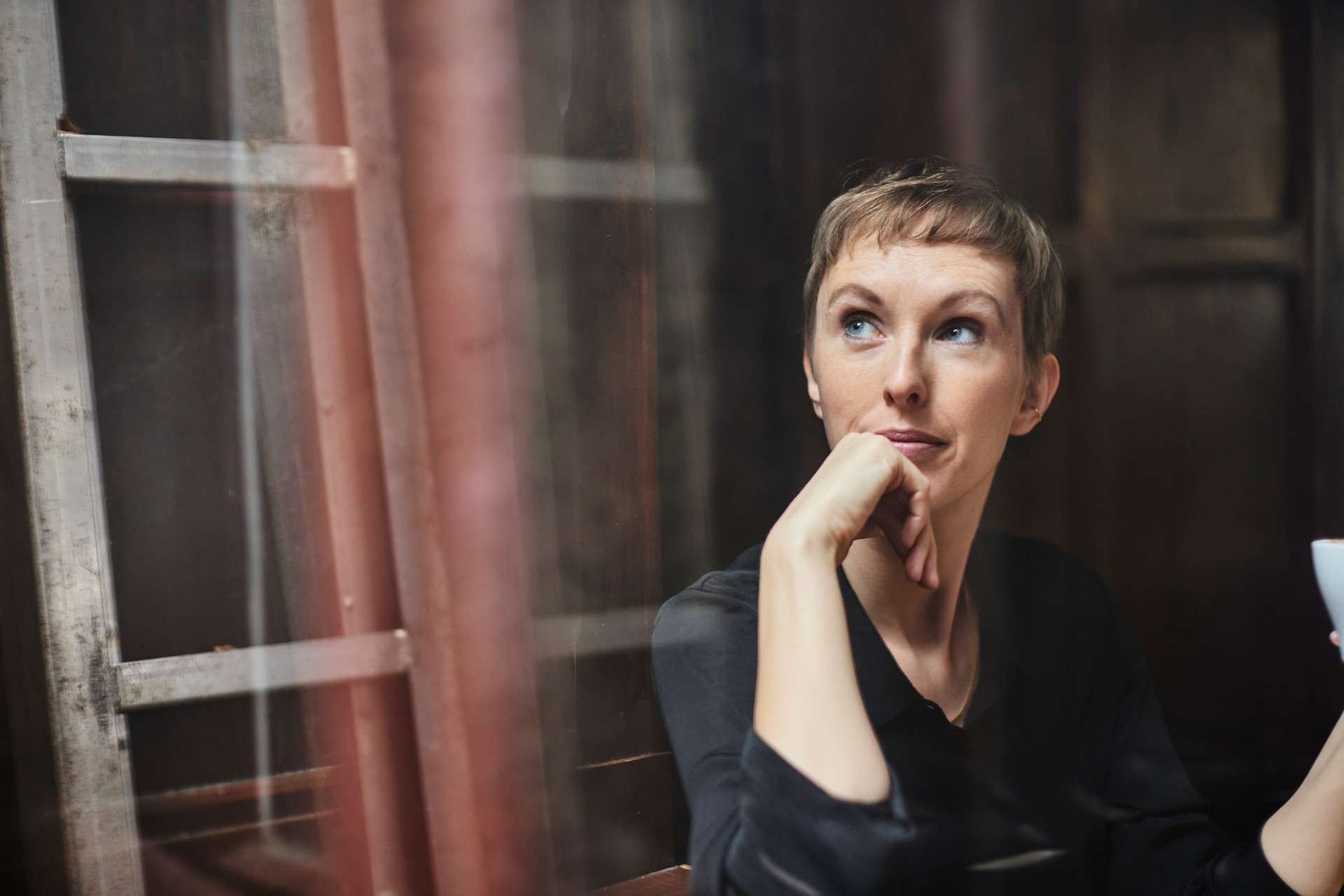 Une personne aux cheveux courts et blonds est assise dans une pièce sombre. Elle porte un haut noir et tient une tasse dans sa main droite. En arrière-plan, on voit une échelle en bois et un mur.