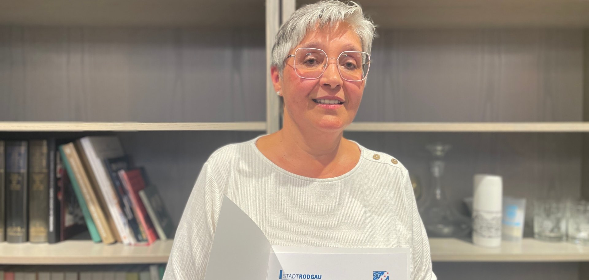 Une personne tient un certificat avec le texte 'Hona Szycharz 40e anniversaire de service'. Des étagères et un vase sont visibles à l'arrière-plan.