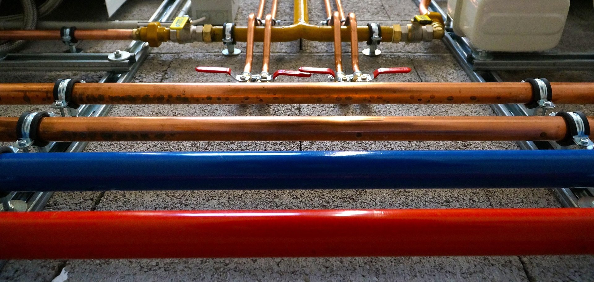 Tuyauterie de différentes couleurs (rouge, bleu, orange) avec vannes et raccords installés sur un mur en béton.