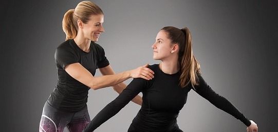 Deux femmes en tenue de sport. La femme de gauche porte un t-shirt noir et un legging avec des motifs violets. La femme de droite porte un haut noir et un legging noir, avec des chaussures roses. La femme de gauche soutient celle de droite, qui se tient dans une pose sportive.