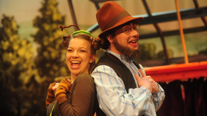 Thater Deux acteurs en costume de théâtre, l'un portant un chapeau et un gilet marron, l'autre un bonnet coloré avec des tresses, se tiennent dos à dos.