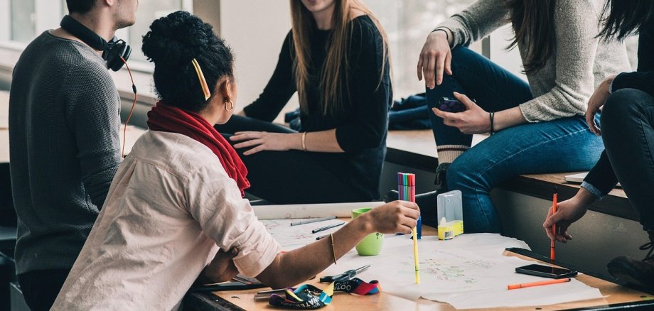 Plusieurs jeunes adultes sont assis autour d'une table dans un café linguistique, discutent et travaillent ensemble sur une grande feuille de papier avec des stylos et des marqueurs.