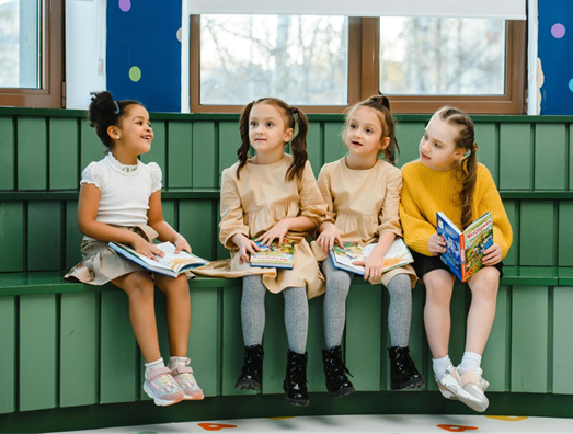 quatre filles aux cheveux longs sont assises sur une estrade et tiennent chacune un livre à la main. Elles se lisent un passage et ont l'air joyeuses.
