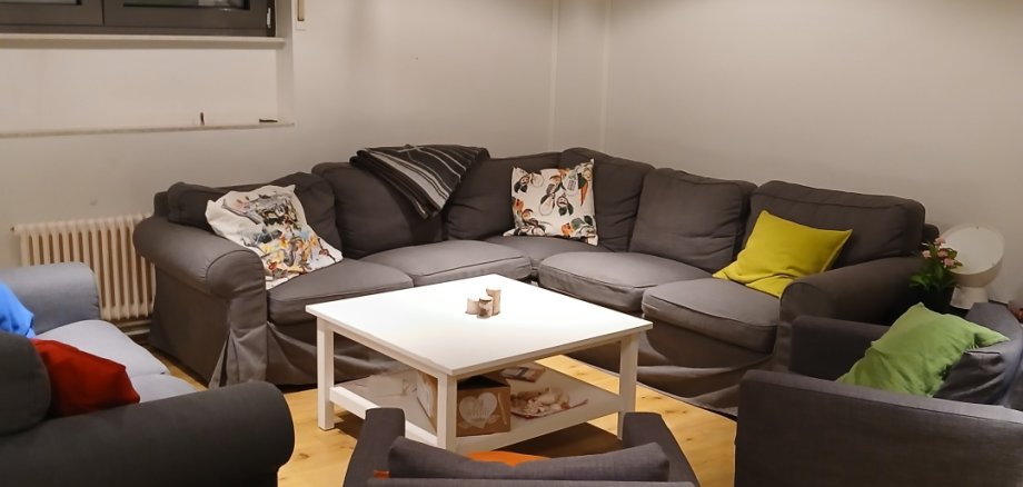 Il y a un canapé d'angle gris et trois fauteuils dans cette pièce. Il y a beaucoup de coussins colorés sur ces derniers et une table blanche à côté.