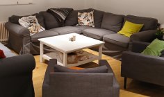 Centre de jeunes Il y a un canapé d'angle gris et trois fauteuils dans cette pièce. Il y a beaucoup de coussins colorés sur ces derniers et une table blanche à côté.