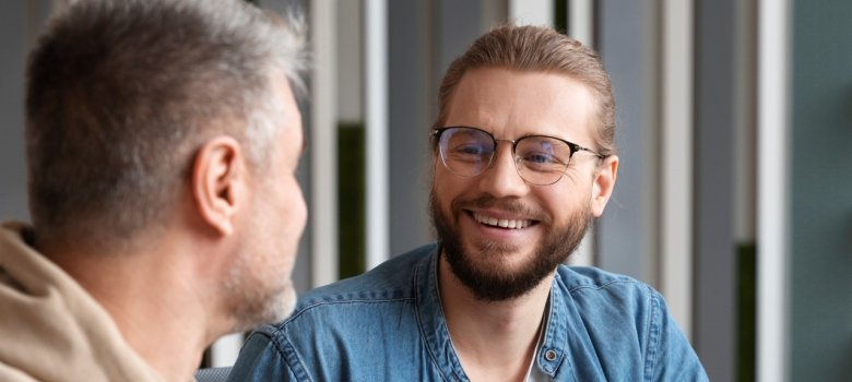 Deux hommes en train de discuter, l'un portant un sweat à capuche beige, l'autre une veste en jean bleu. Ils se sourient