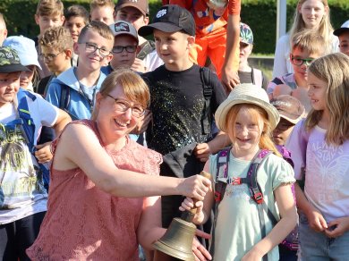 Ouverture des jeux de vacances Mme Martin sonne une cloche pour l'ouverture des jeux de vacances, entourée d'enfants avec des sacs à dos à l'extérieur
