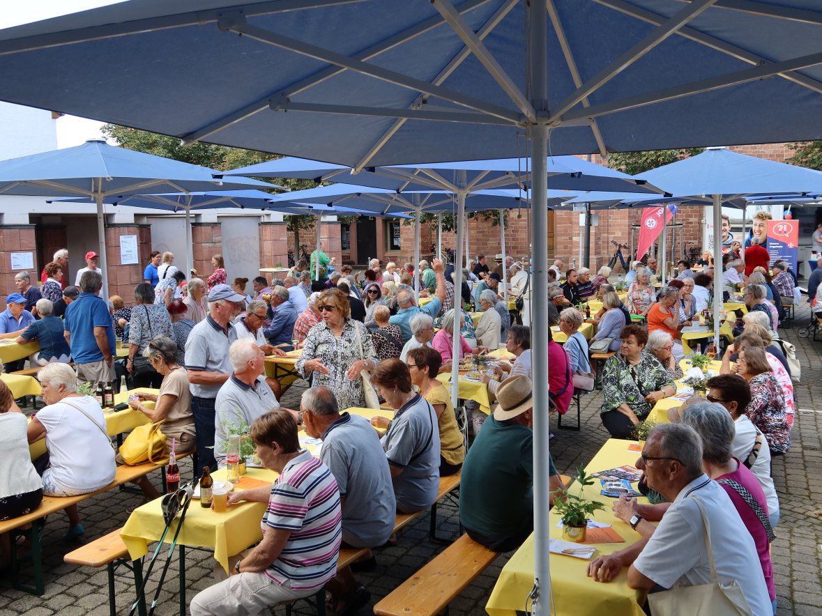 De nombreuses personnes âgées lors d'un événement De nombreuses personnes âgées sont assises sous des parasols bleus à de longues tables en bois recouvertes de nappes jaunes lors d'un événement en plein air.