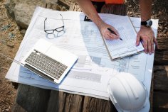 Chantier sur une table se trouve un plan de construction avec un ordinateur portable, des lunettes de protection et un casque. Un homme se tient à côté de la table et montre les documents.