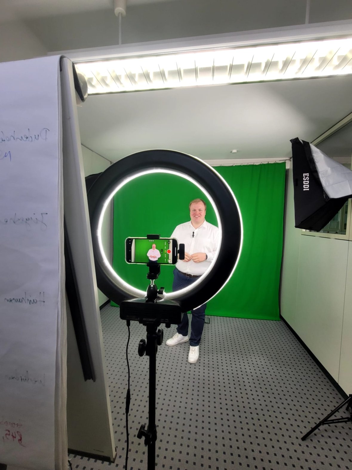 Le maire Max Breitenbach, vêtu d'une chemise blanche et d'un pantalon sombre, se tient devant un fond vert, photographié par un éclairage annulaire avec un smartphone sur trépied, entouré d'un éclairage de studio.