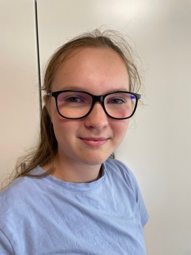 Jeune femme aux cheveux châtain clair, portant des lunettes noires et un t-shirt bleu clair, devant un mur blanc