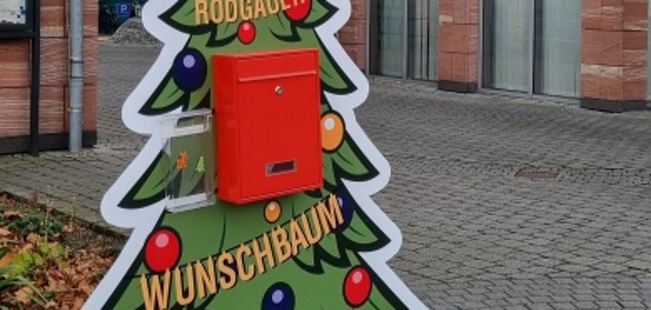 Un panneau coloré en forme d'arbre de Noël avec une boîte aux lettres rouge, portant l'inscription 'RODGAUER WUNSCHBAUM', se trouve sur une place pavée devant le bâtiment de la mairie.