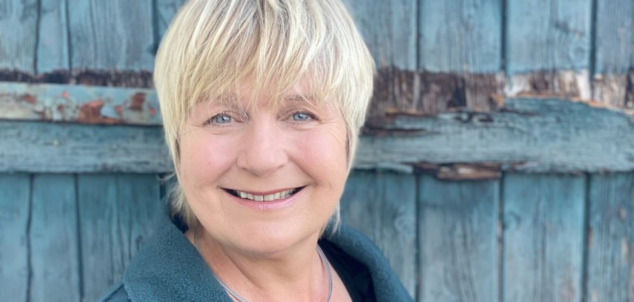 Maja Nielsen, con i capelli corti e biondi, indossa un cappotto verde scuro e una collana con un ciondolo rotondo davanti a una porta di legno blu, danneggiata dalle intemperie.