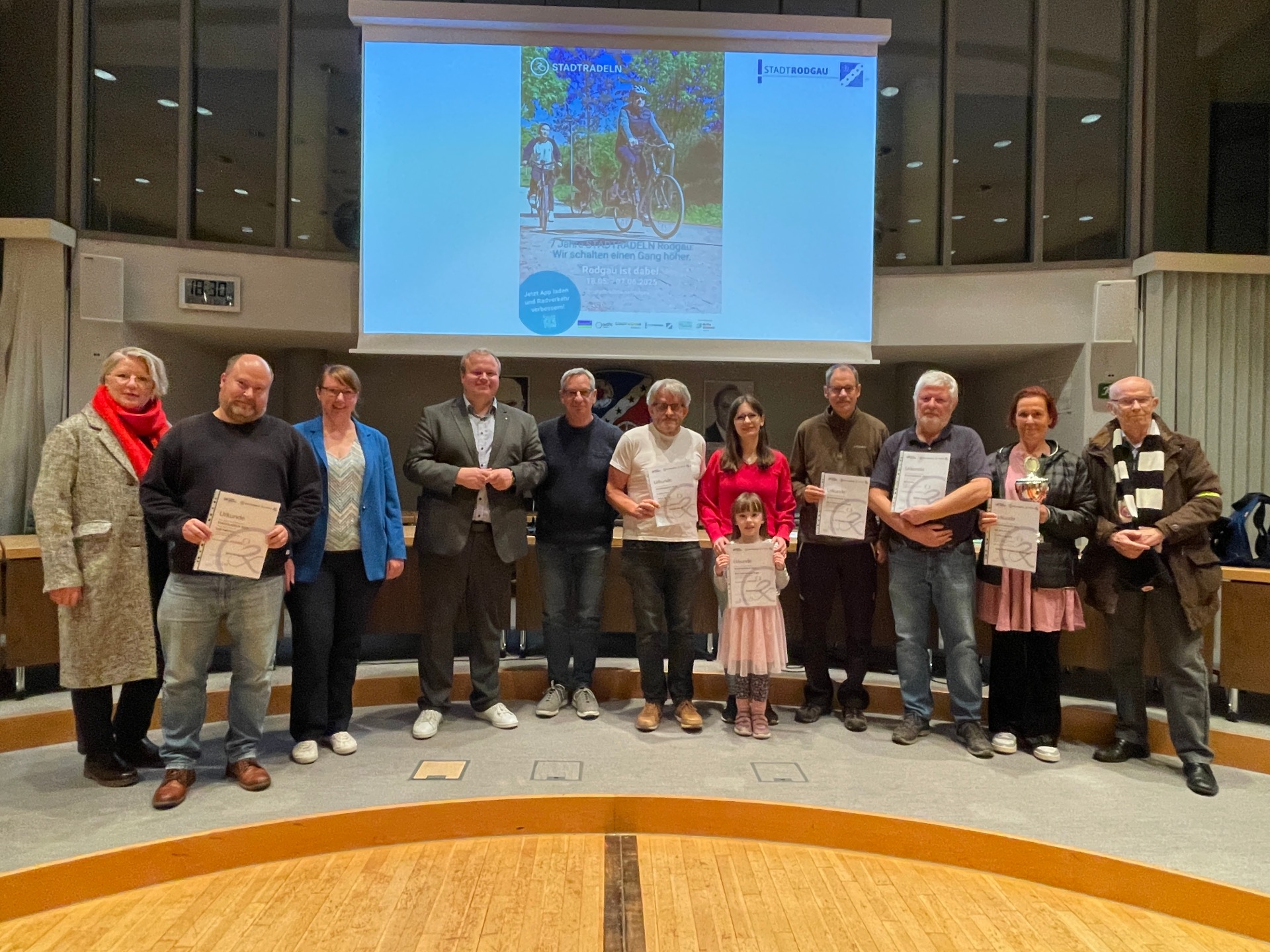 Gruppo di dodici persone in piedi su un palco, alcune con in mano dei certificati, sullo sfondo l'immagine di un proiettore con il motivo di una bicicletta e un testo sull'evento conclusivo di CITY CYCLING.