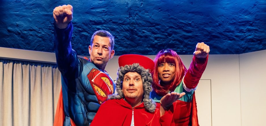 Tre persone su un palco: una con mantello rosso e parrucca grigia, due in costume da supereroe con mantello rosso, ognuna delle quali tiene un pugno davanti a sé.