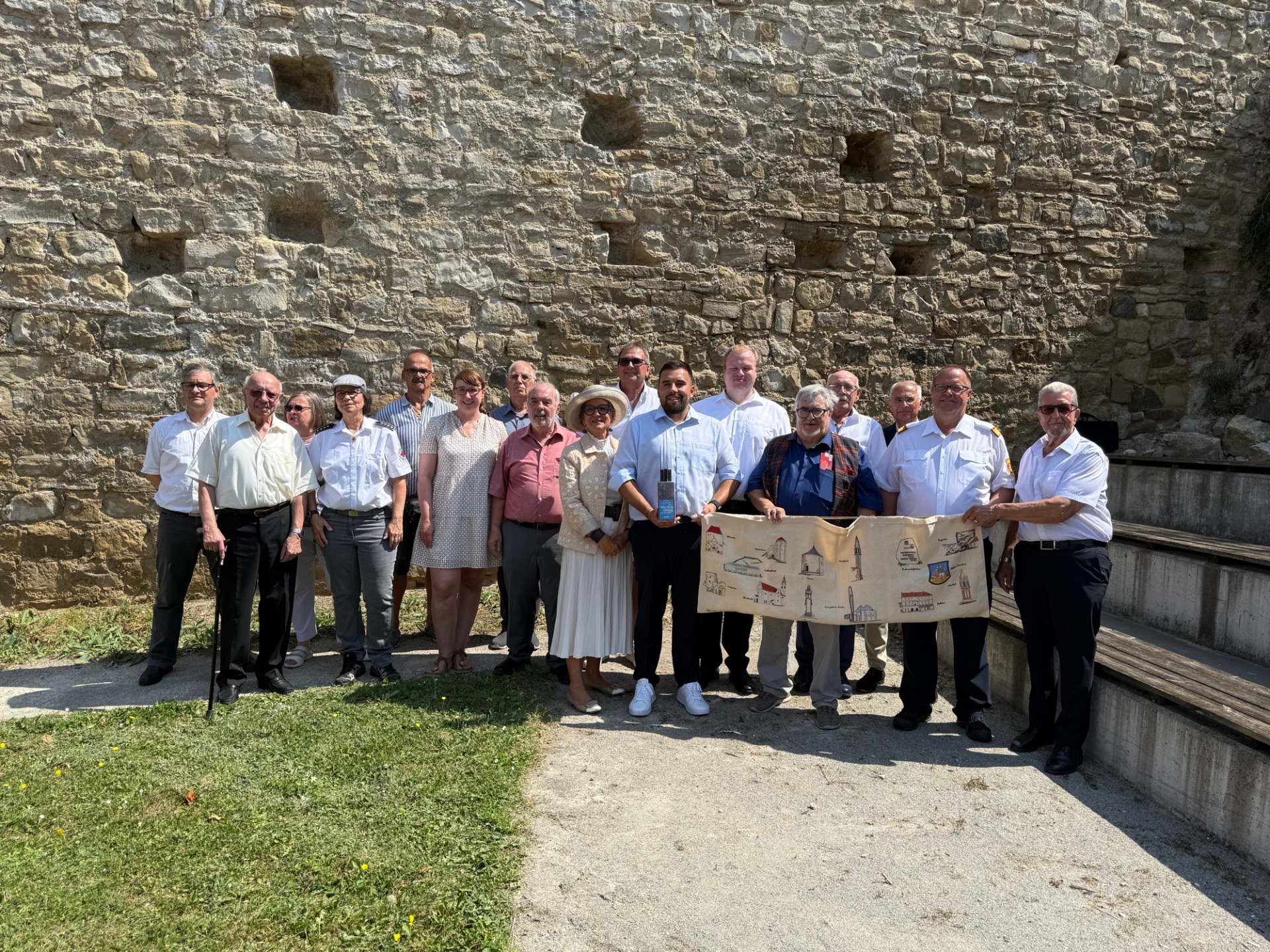Un gruppo di 14 persone si trova davanti a un muro di pietra. Alcune tengono in mano un grande foglio di carta piegato con dei disegni. L'abbigliamento varia da formale a casual.