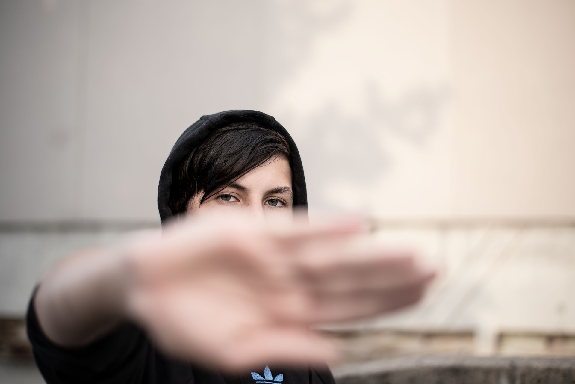 Donna con giacca nera con cappuccio tiene il palmo della mano verso la telecamera, sfondo sfocato con muro e graffiti