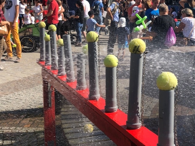 Una stazione idrica rossa con diversi tubi grigi con palline da tennis su catene. Sullo sfondo ci sono molte persone che partecipano a una festa.