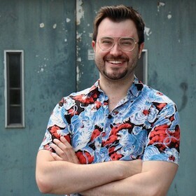 Uomo Uomo a braccia conserte e con un grande sorriso che indossa una camicia a maniche corte con un motivo floreale blu e rosso davanti a un muro grigio con due finestre strette