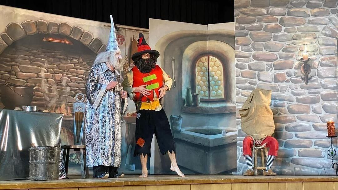 Scena teatrale con tre persone in costume: un mago con lunga barba bianca e cappello a punta, una figura con camicia rossa, cappello e barba neri e una persona seduta con un sacco in testa in pantaloni rossi.