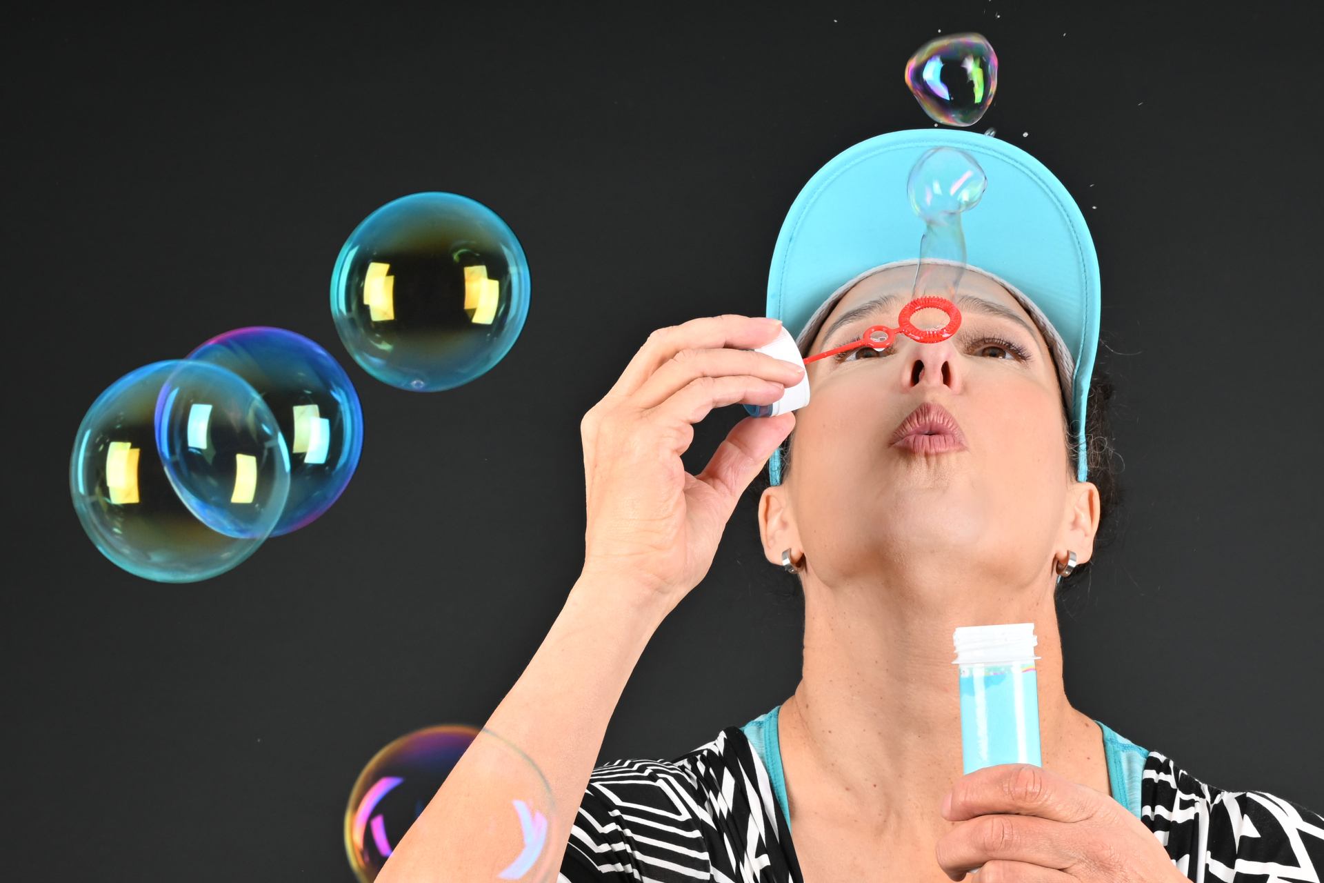 Stefanie Kerker 2023 Donna che soffia bolle di sapone su sfondo nero, con top a righe e copricapo blu chiaro