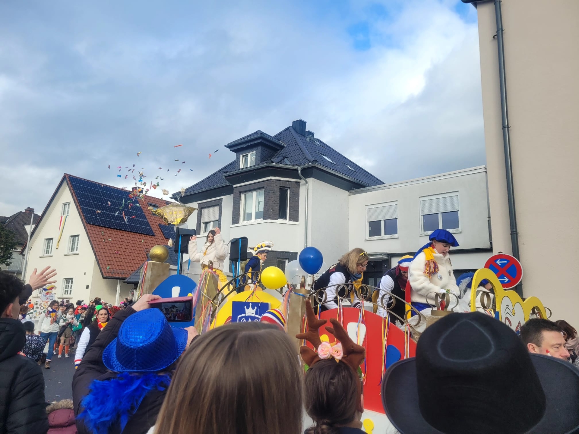 La principessa e il principe dei bambini in costume festivo su un carro colorato alla sfilata di carnevale davanti alle case, circondati da spettatori festanti con cappelli e palloncini