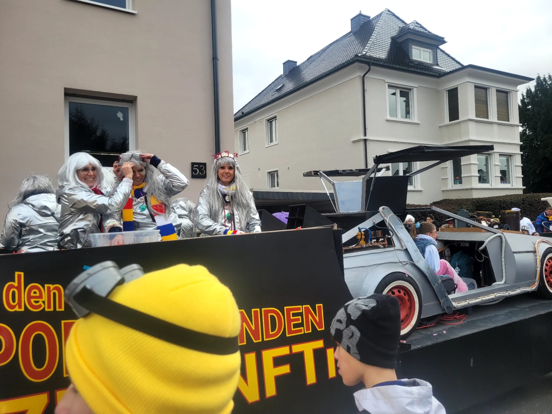 Sfilata di Carnevale 2026 con un carro che mostra una replica della DeLorean di "Ritorno al futuro", persone con tute spaziali argentate e un bambino con un berretto giallo e nero in primo piano.