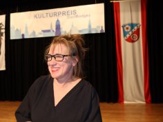 Pirkko Cremer con top nero davanti a una tenda nera, sopra uno striscione con la scritta "KULTURPREIS Stadt Rodgau" e una bandiera rossa e bianca con stemma appesa a destra.