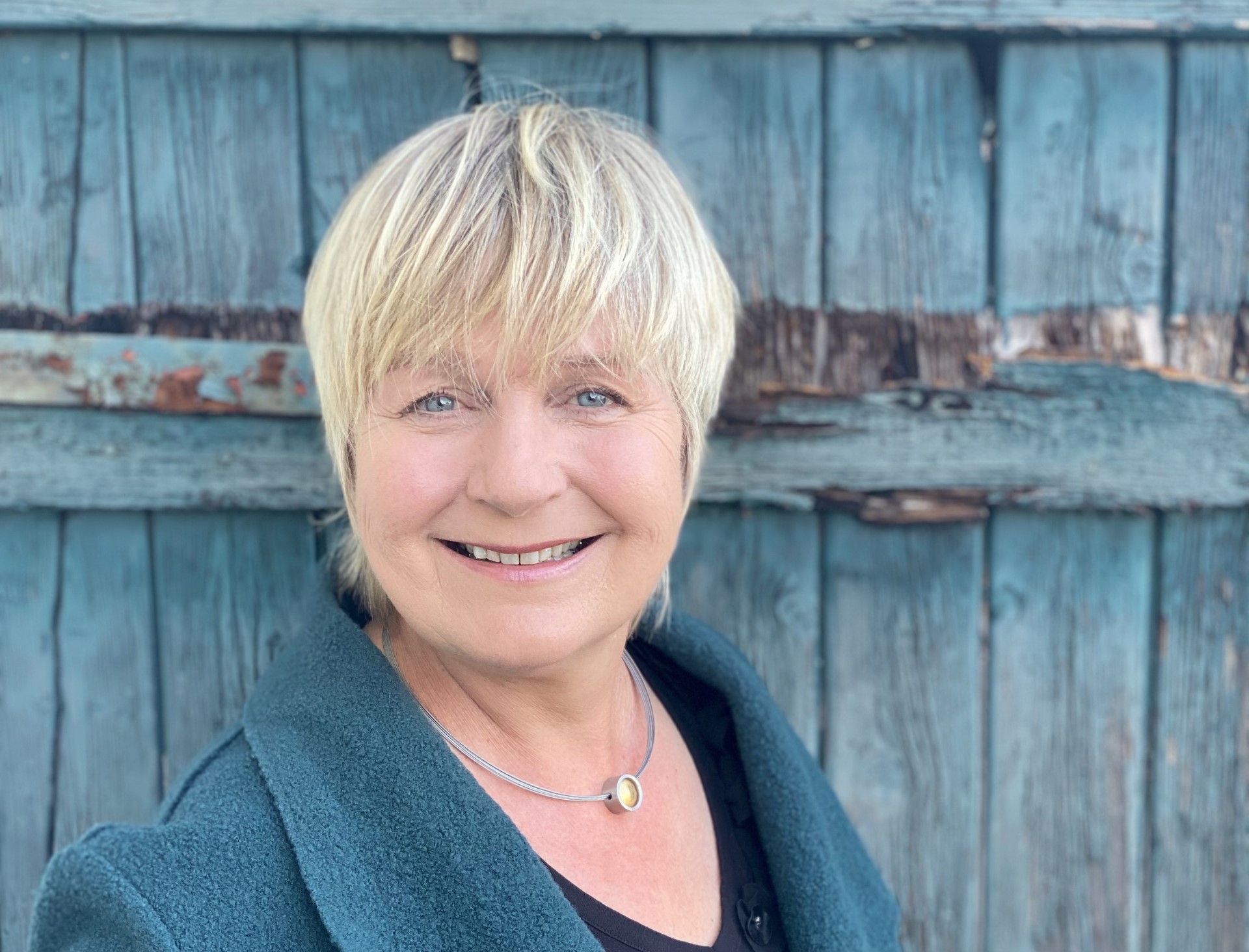 Maja Nielsen, z krótkimi blond włosami, nosi ciemnozielony płaszcz i naszyjnik z okrągłym wisiorkiem przed zwietrzałymi niebieskimi drewnianymi drzwiami.