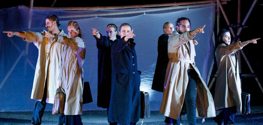 Teatr uliczny pod drzewami Scena jest ciemnoniebieska. Sześć osób w długich, jasnych płaszczach stoi w rzędzie i wskazuje palcami w różnych kierunkach. Niektórzy trzymają w rękach torby.