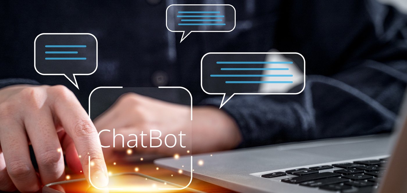 Ręka dotyka smartfona, obok widać laptopa. Wirtualnie widać dymek z terminem "chatbot".