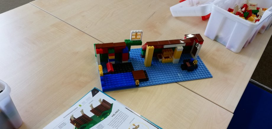 Wielka akcja budowlana dla dzieci w bibliotece miejskiej w Nieder-Roden Na stole stoi częściowo zbudowany model z klocków Lego na niebieskiej podstawie, obok otwartej instrukcji montażu oraz dwóch białych pojemników z klockami Lego.