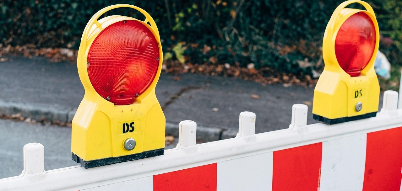 Dwa żółte światła ostrzegawcze z czerwonym światłem na białej barierze z czerwonymi i białymi paskami. W tle widoczne są zielone rośliny.