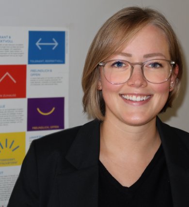 Pani Hannig ma na sobie czarną marynarkę, czarną koszulę i okulary i stoi przed ścianą z kolorowym plakatem