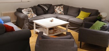 W tym pokoju znajduje się szara narożna sofa i trzy fotele. Znajduje się na nich wiele kolorowych poduszek i biały stolik.