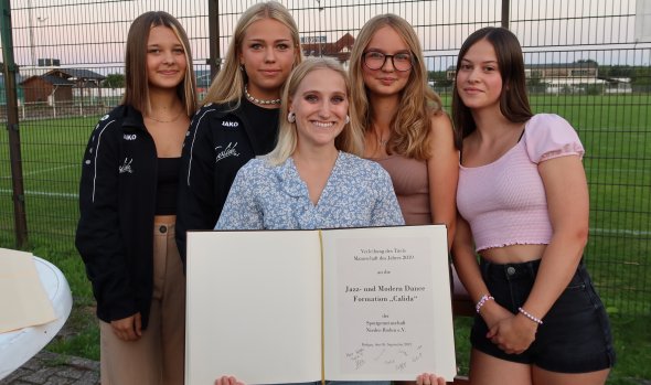Pięć młodych kobiet stoi na zewnątrz przed płotem, jedna z nich trzyma złotą księgę z nagrodą za taniec jazzowy i nowoczesny od formacji Calida