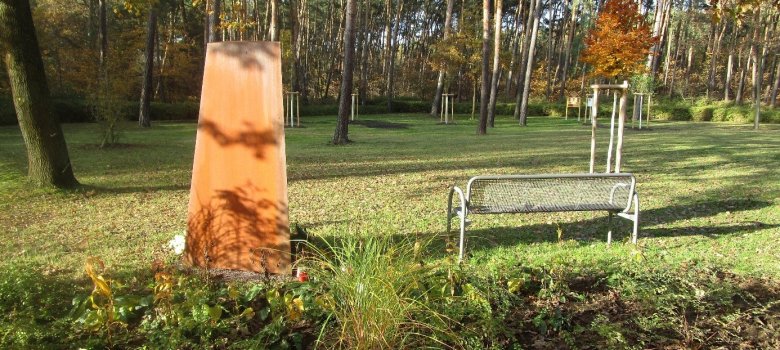 Widok skierowany jest na trawnik. Po lewej stela z zardzewiałej stali, po prawej ławka. W tle widać sosny.