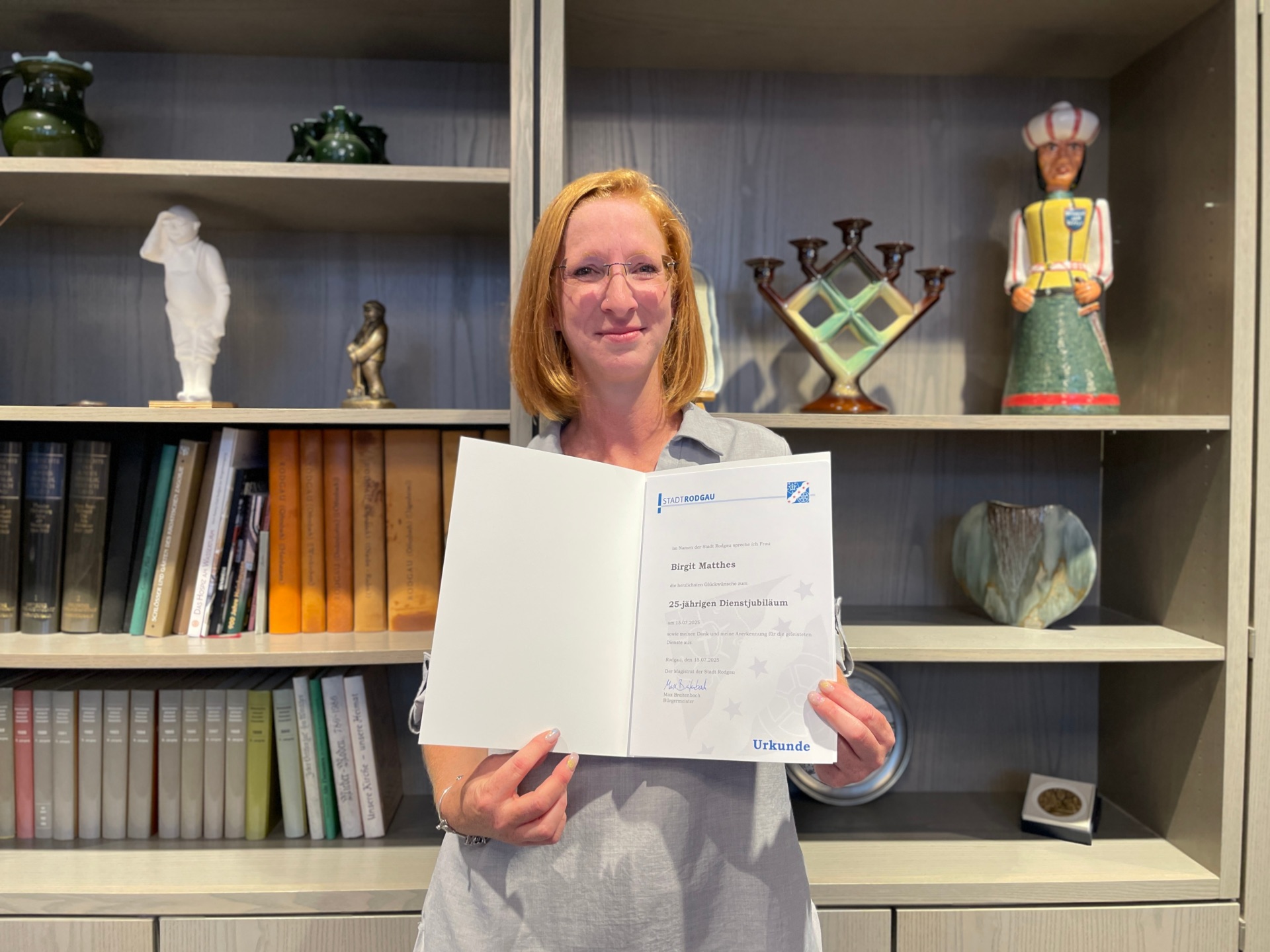 Birgit Mattes segura um certificado com a inscrição "25 anos de serviço". Em segundo plano, encontram-se livros, uma estátua e uma vitrina decorativa.