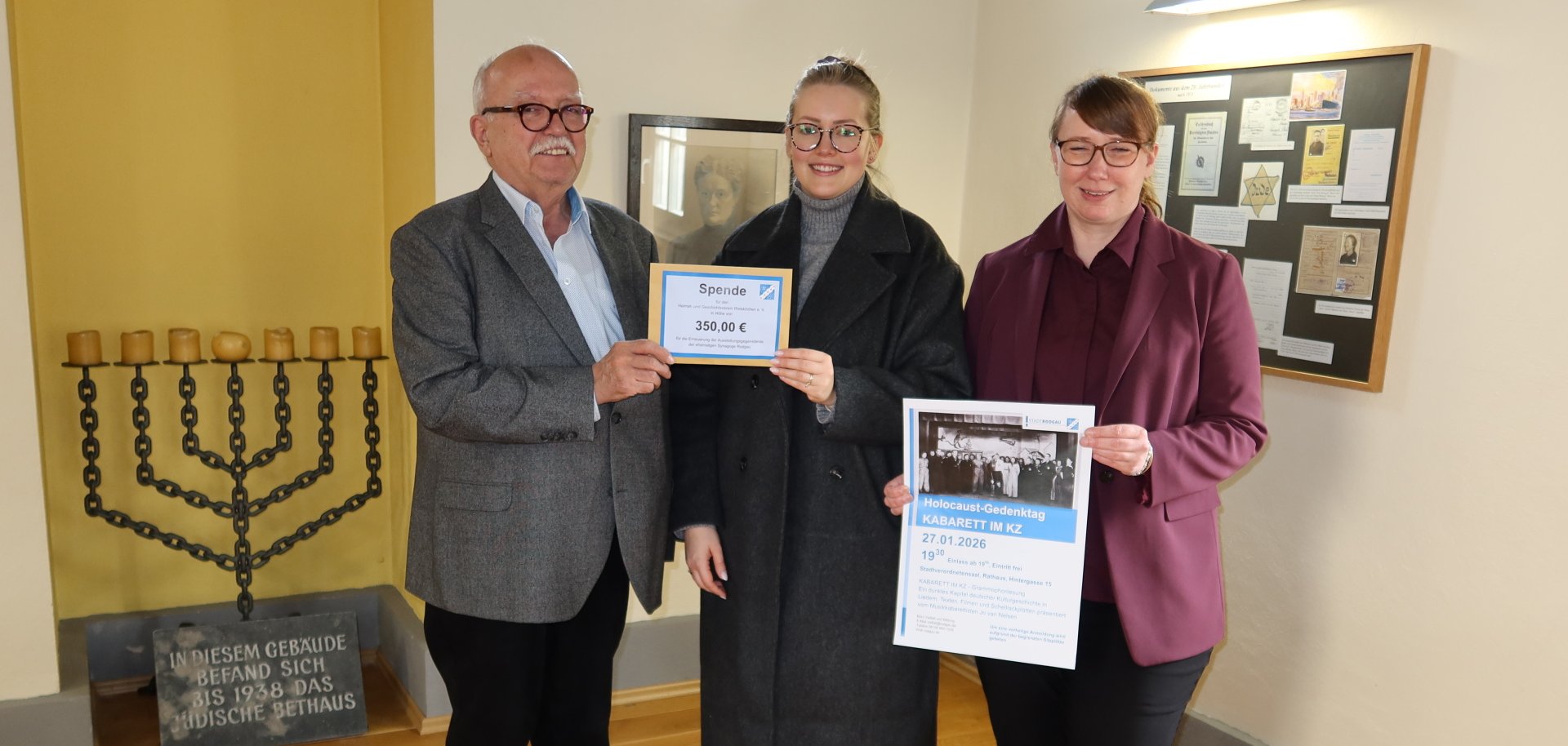 Três pessoas estão de pé numa sala com um candelabro de menorá e uma placa comemorativa, segurando um cartaz que diz "Donativo 350,00 euros" e um cartaz do evento comemorativo do Holocausto.