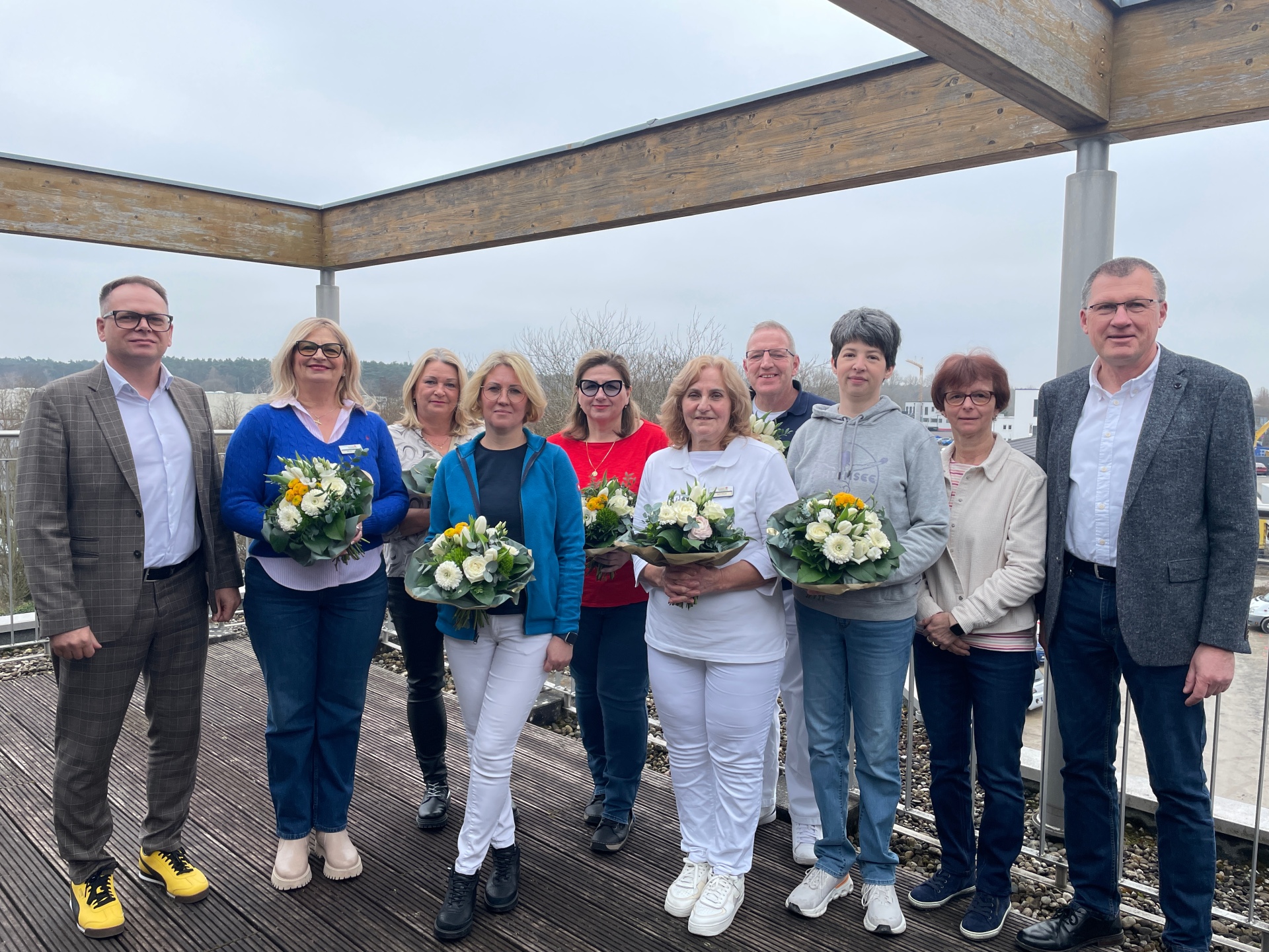 Grupo de onze pessoas de pé num terraço de madeira em frente a uma paisagem, várias mulheres segurando ramos de flores, dois homens de pé ao lado.