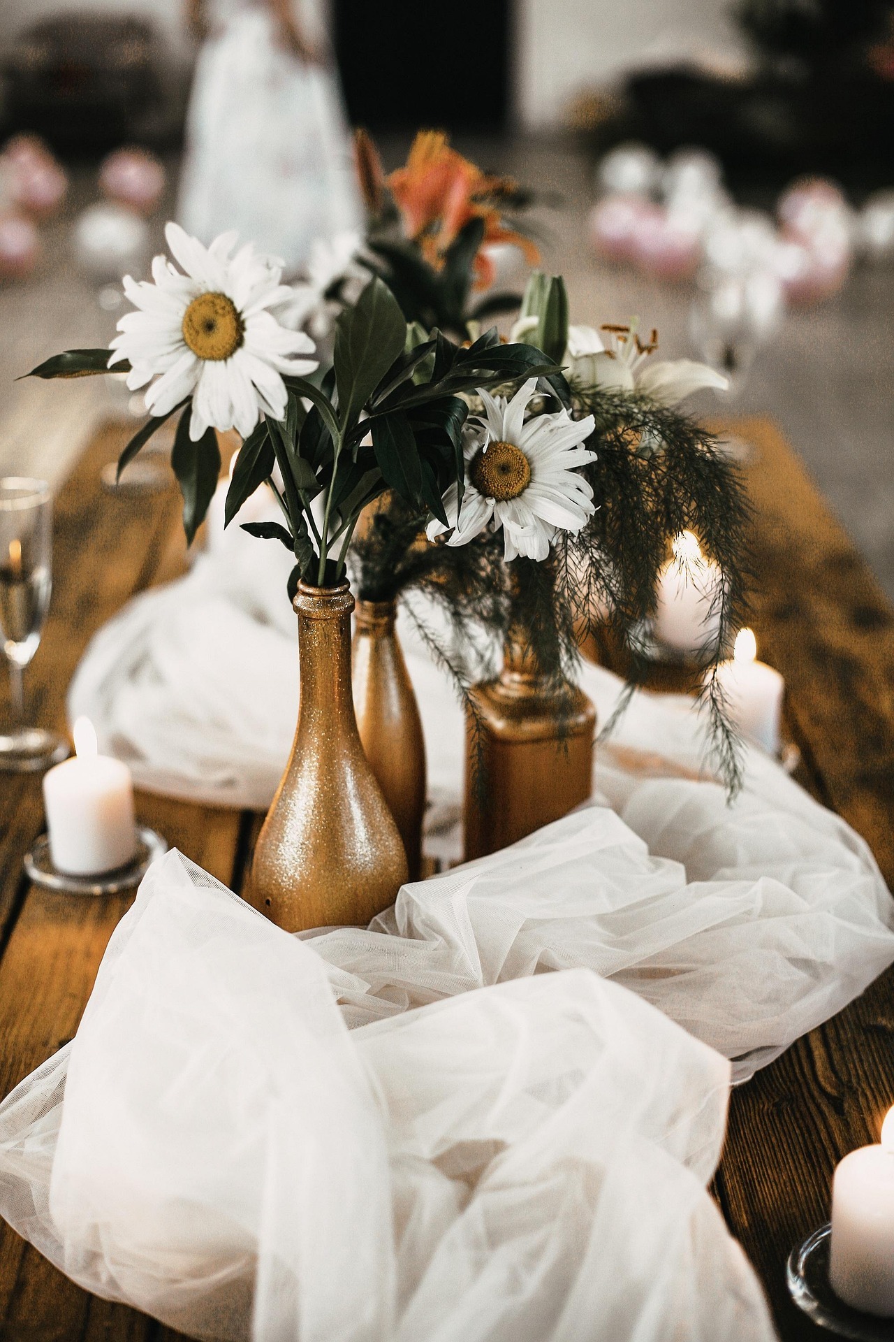 Decoração de mesa com margaridas brancas em vasos dourados, velas brancas e um tecido branco solto sobre uma mesa de madeira, no fundo esbatido, outras mesas com decoração semelhante