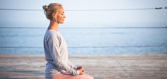 vhs Mindfulness Uma pessoa está sentada em posição de lótus num convés de madeira junto à água. Está vestida com um top cinzento e umas leggings cor-de-rosa. O fundo mostra água calma e um céu limpo.