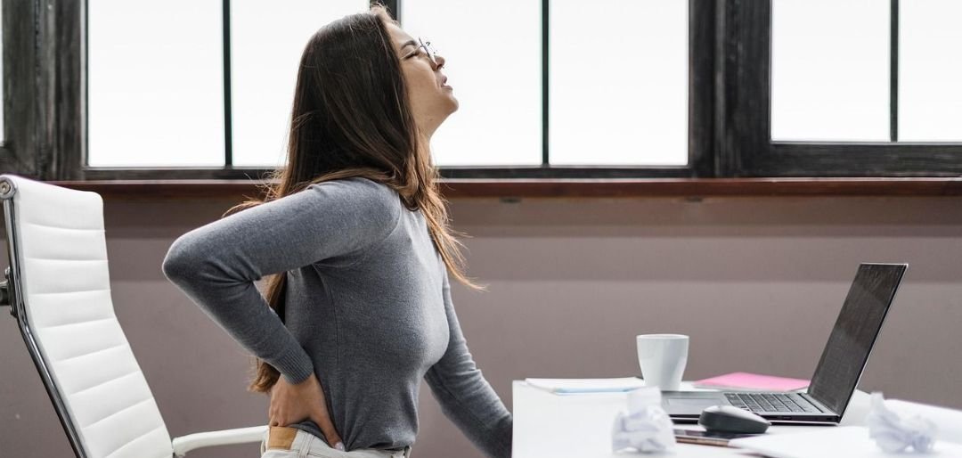 Duas mulheres com roupa de desporto. A mulher da esquerda veste uma T-shirt preta e umas leggings com um padrão roxo. A mulher da direita veste um top preto e umas leggings pretas, com sapatos cor-de-rosa. A mulher da esquerda está a apoiar a mulher da direita, que está numa pose desportiva.