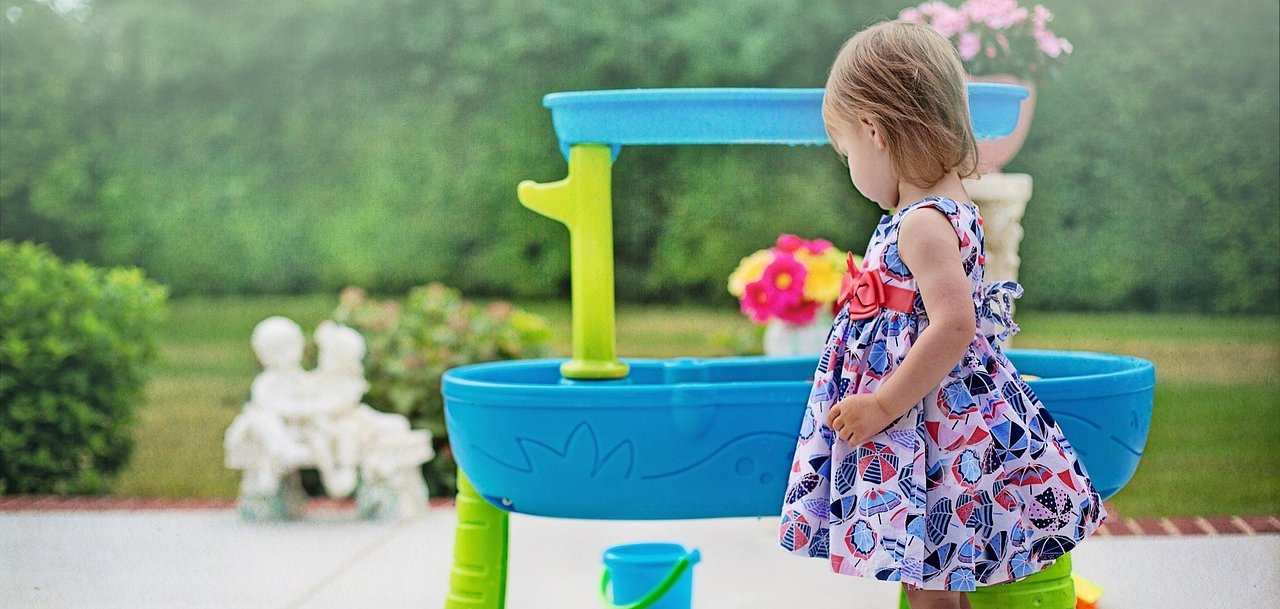 Menina com vestido estampado e sandálias em frente a uma mesa de jogo azul e verde com balde no jardim