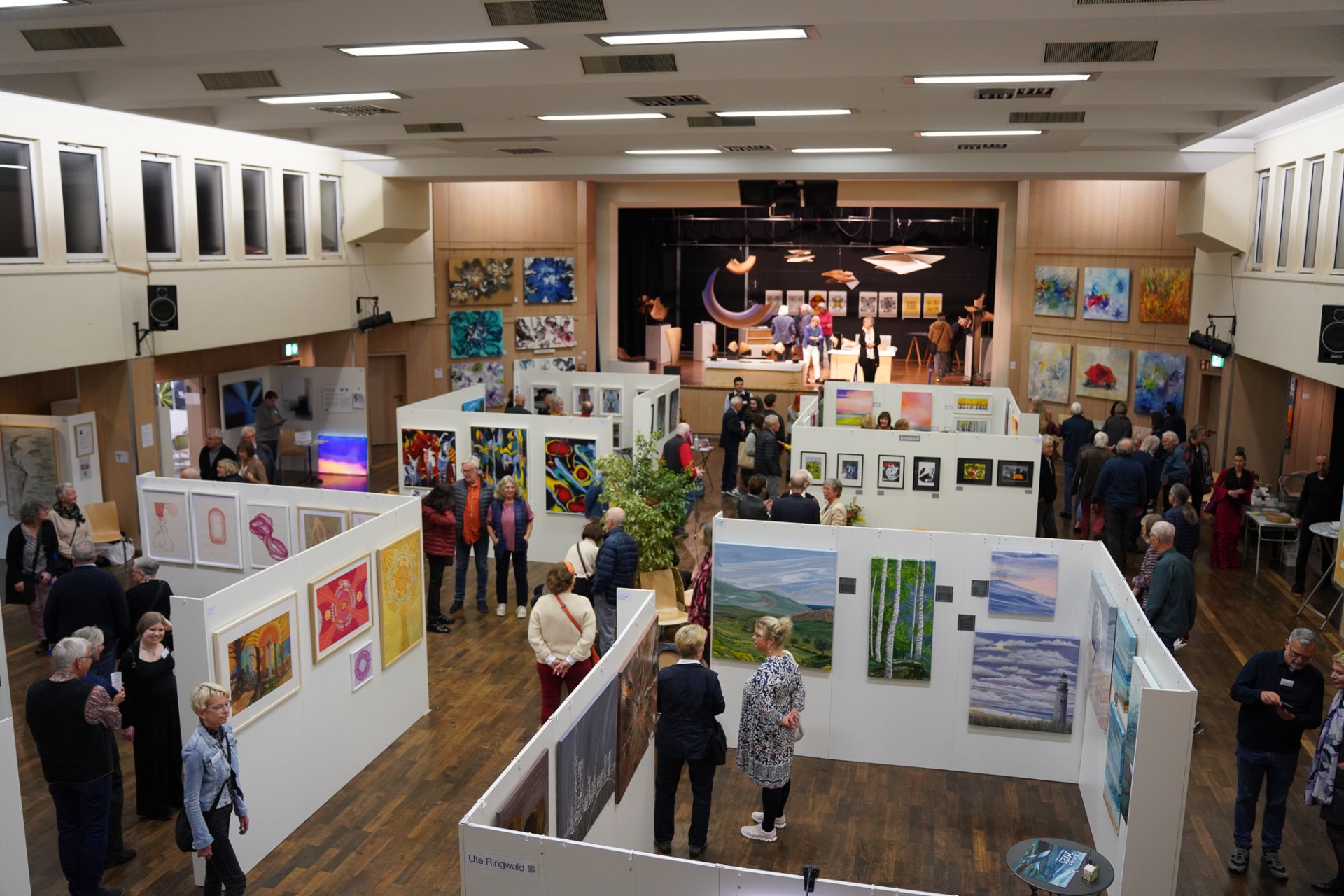 Os visitantes vêem pinturas e esculturas numa sala de exposições na RodgauArt, com obras de arte em paredes brancas e um palco ao fundo