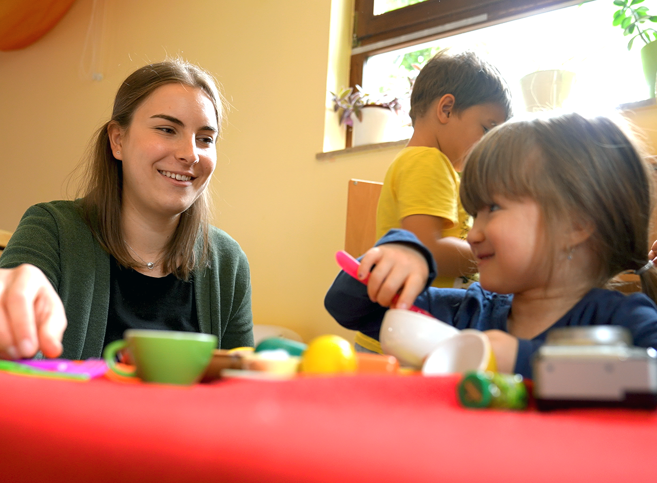 A professora (à esquerda) brinca com duas crianças (à direita) numa cozinha de brincar. O interior de uma creche pode ser visto ao fundo.