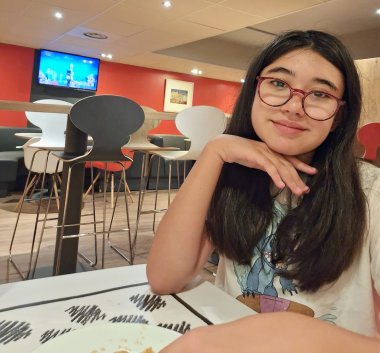 Uma rapariga com cabelo comprido e escuro e óculos vermelhos está sentada à mesa de um café, com o queixo apoiado na mão