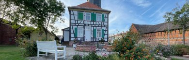 Casa do Reitor Geissler com jardim Rektor-Geißler-Haus em Dudenhofen, uma casa histórica em enxaimel com portadas verdes e brancas e uma torre, rodeada por um jardim com flores e um banco branco.
